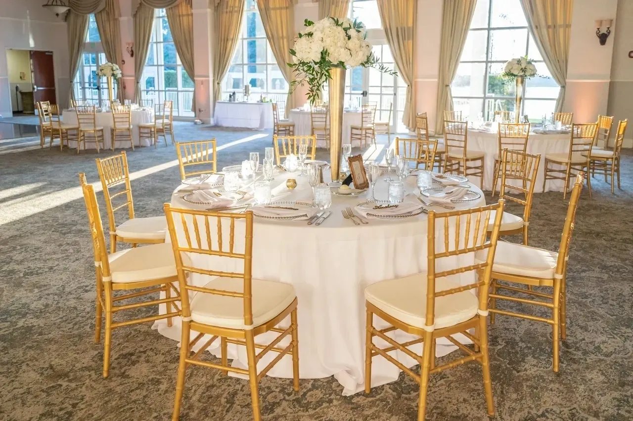 A round table with white linens and gold chairs.